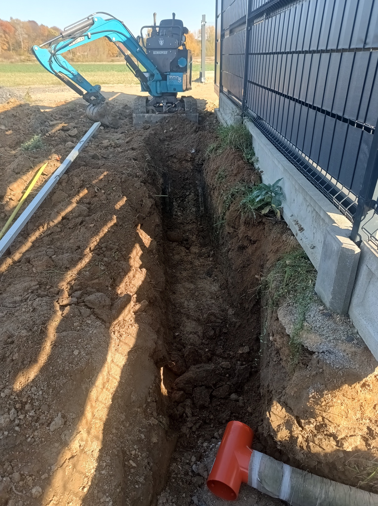 Wąski wykop pod ogrodzeniem z użyciem mini koparki w Lwówku Śląskim. Widoczny poziomica i rura. Praca ziemna w trakcie realizacji, oświetlona słońcem.