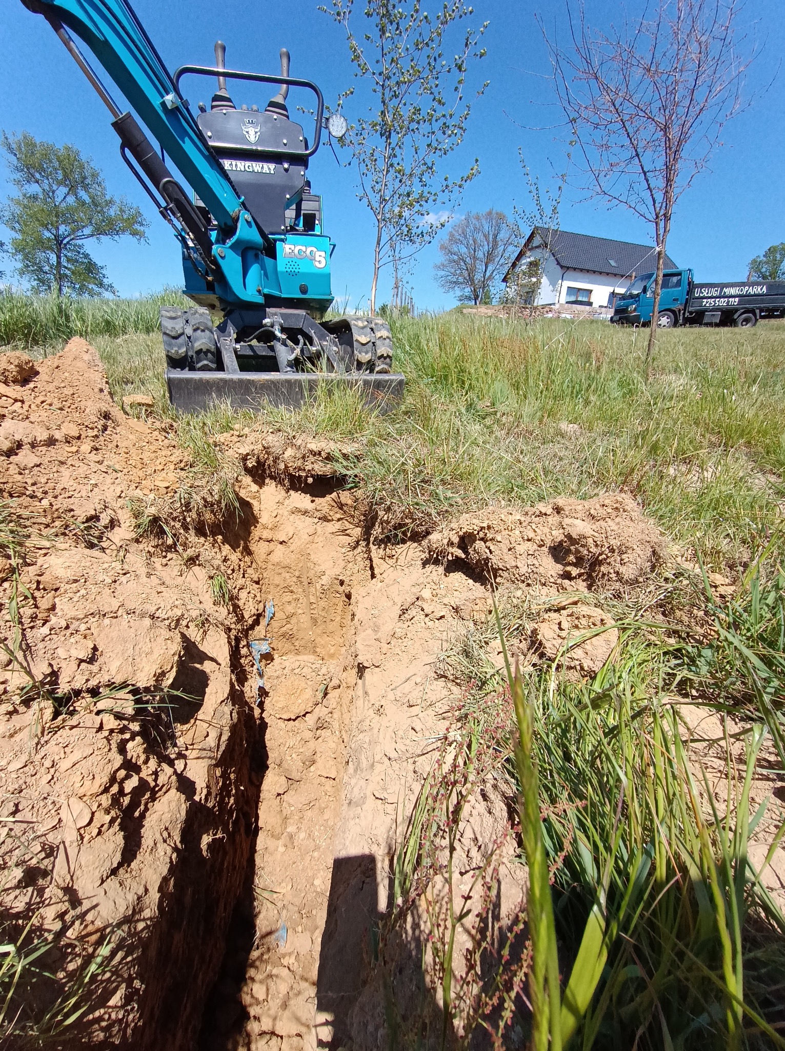 Wąski wykop w ziemi wykonany minikoparką Kingway ECO5. W tle dom jednorodzinny i samochód dostawczy z napisem 'Usługi Minikoparką'.