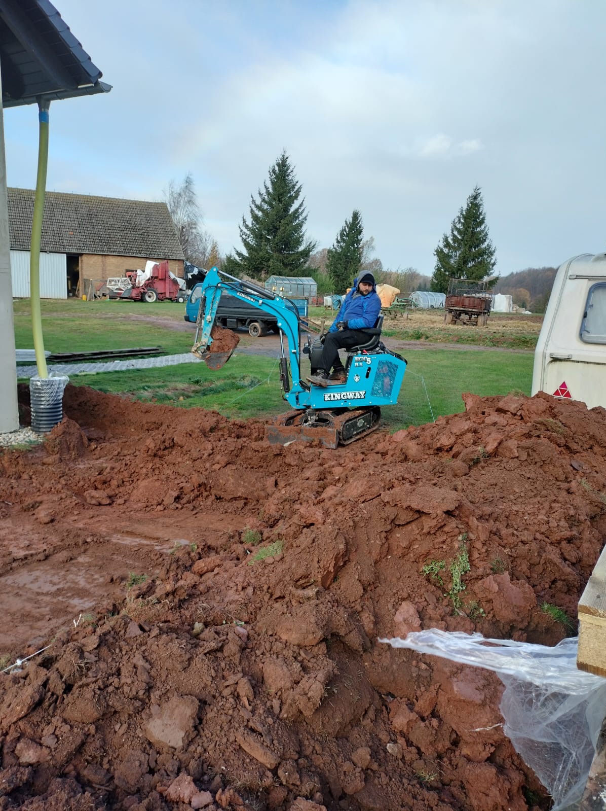 Niebieska mini koparka Kingway z operatorem w trakcie prac ziemnych na działce. W tle budynek gospodarczy i drzewa iglaste.