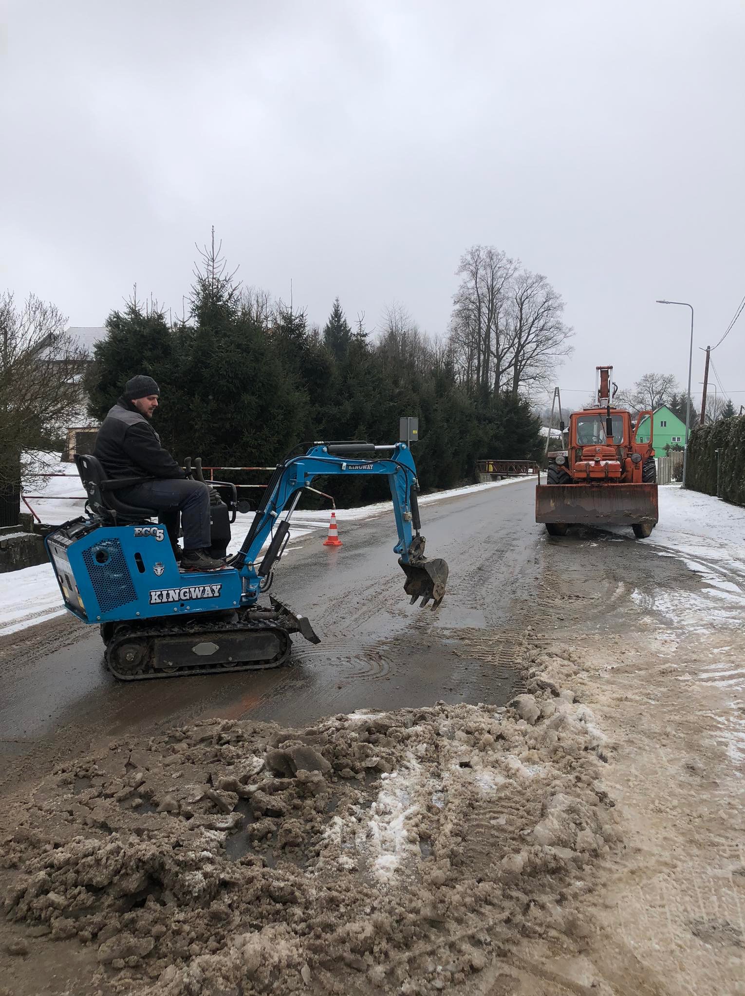Niebieska mini koparka Kingway usuwa błoto pośniegowe z drogi. W tle pomarańczowy traktor z pługiem. Praca w pochmurny dzień.