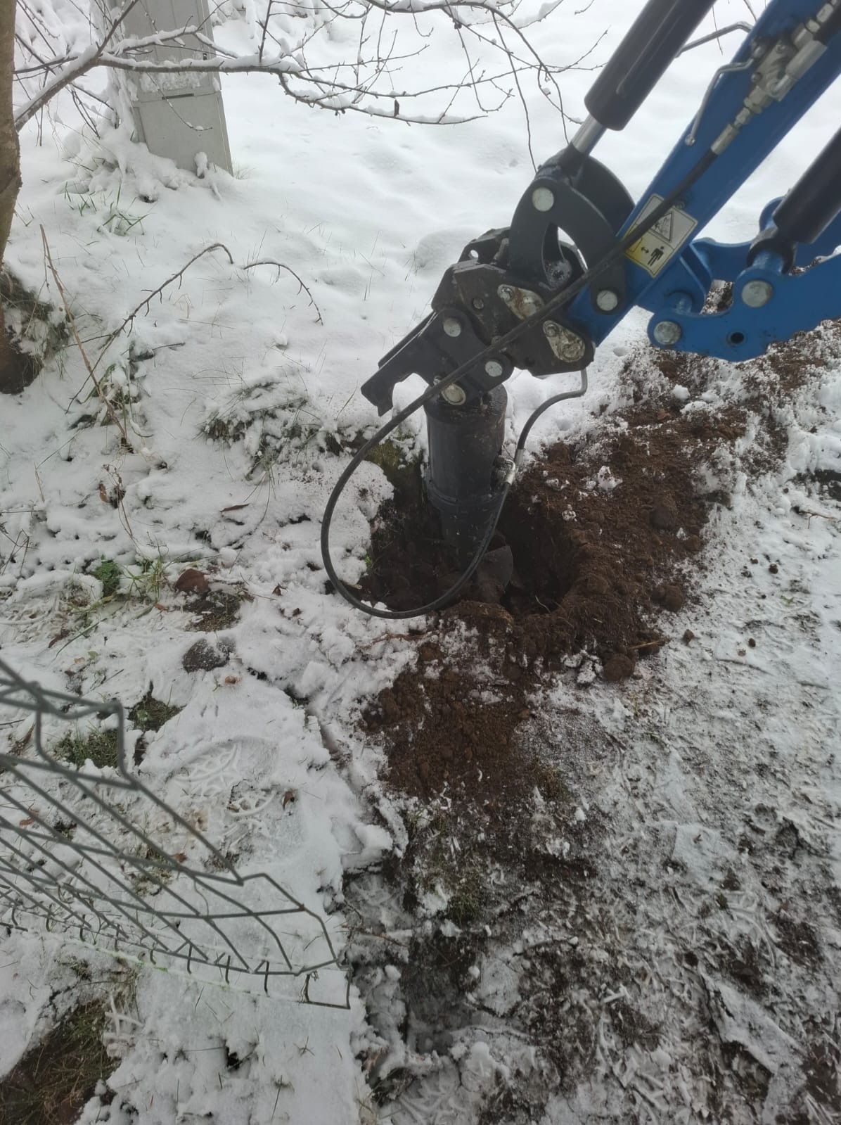 Wiercenie otworu w ziemi w zimowych warunkach za pomocą wiertnicy hydraulicznej. Śnieg leży na ziemi, a w tle widoczny fragment ogrodzenia.