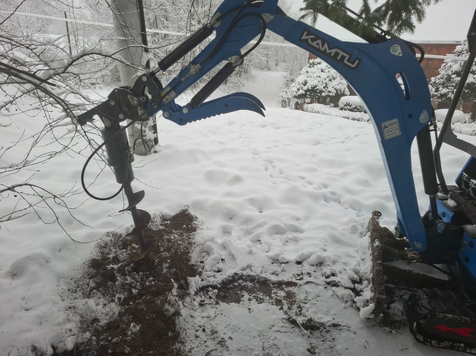 Niebieska wiertnica KAMTU w śniegu wierci otwór w ziemi. W tle drzewa i krzewy pokryte śniegiem. Ślady na śniegu. Zimowy krajobraz.