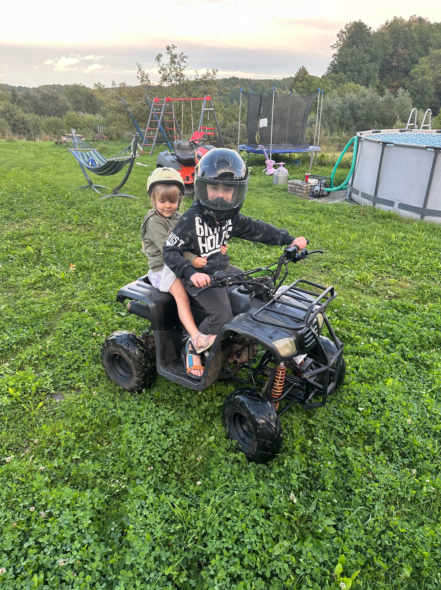 Dzieci w kaskach na czarnym quadzie na tle zielonego trawnika z basenem, trampoliną i huśtawką. Letni dzień, rekreacja na świeżym powietrzu.