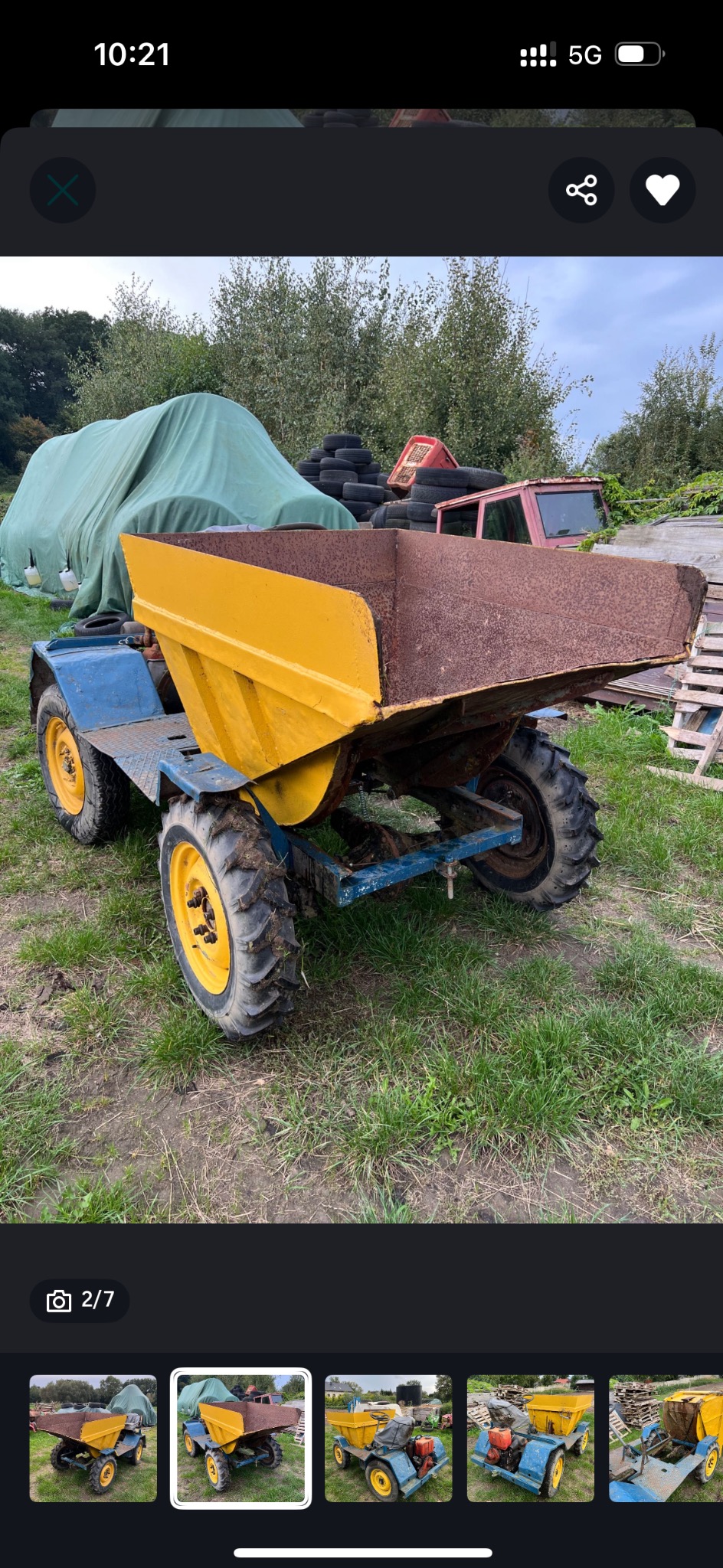 Żółto-niebieska wywrotka na trawiastym terenie, z widocznymi oponami z bieżnikiem i rdzawą skrzynią ładunkową. W tle opony i przykryty plandeką obiekt.