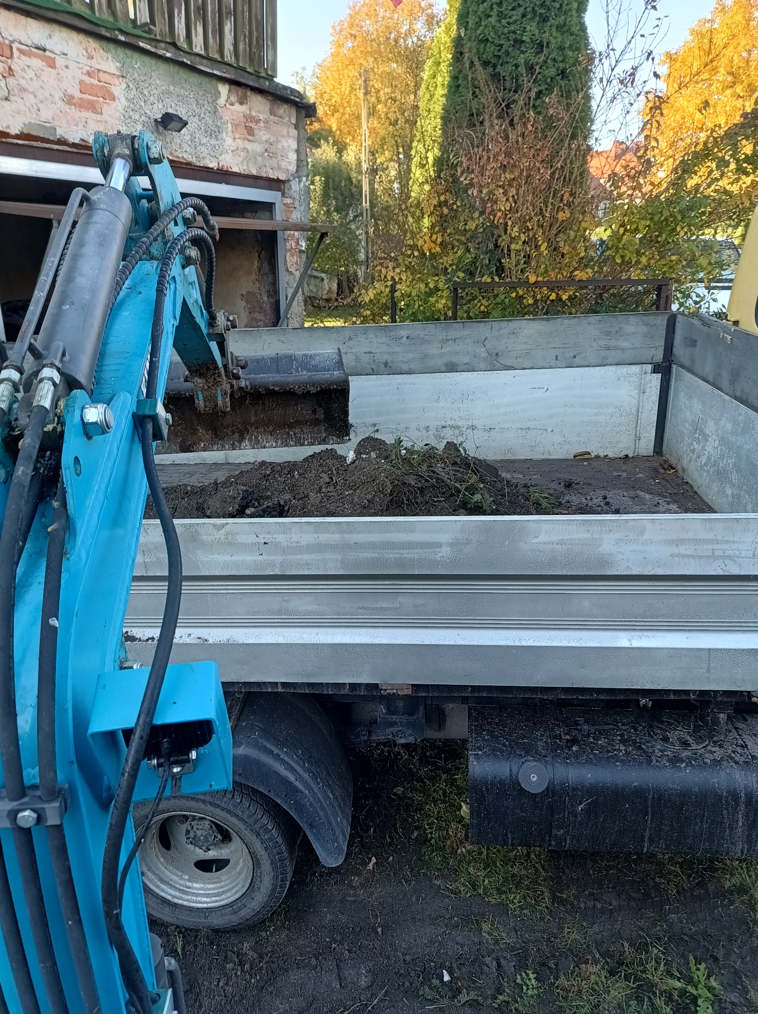 Niebieska mini koparka załadowuje ziemię z wykopu do skrzyni ładunkowej samochodu na tle budynku i drzew w jesiennych barwach.
