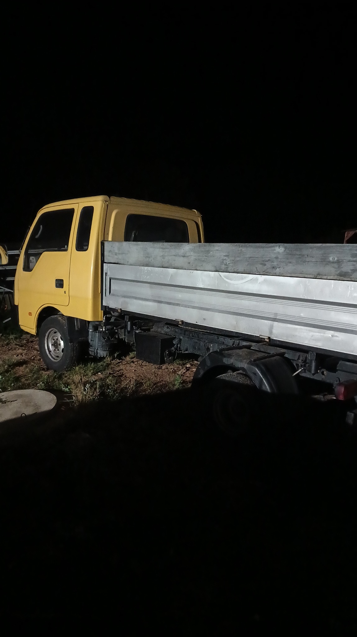 Żółty samochód ciężarowy z otwartą skrzynią ładunkową, zaparkowany na ziemi w nocy. Widoczne opony i elementy podwozia. Słabe oświetlenie.