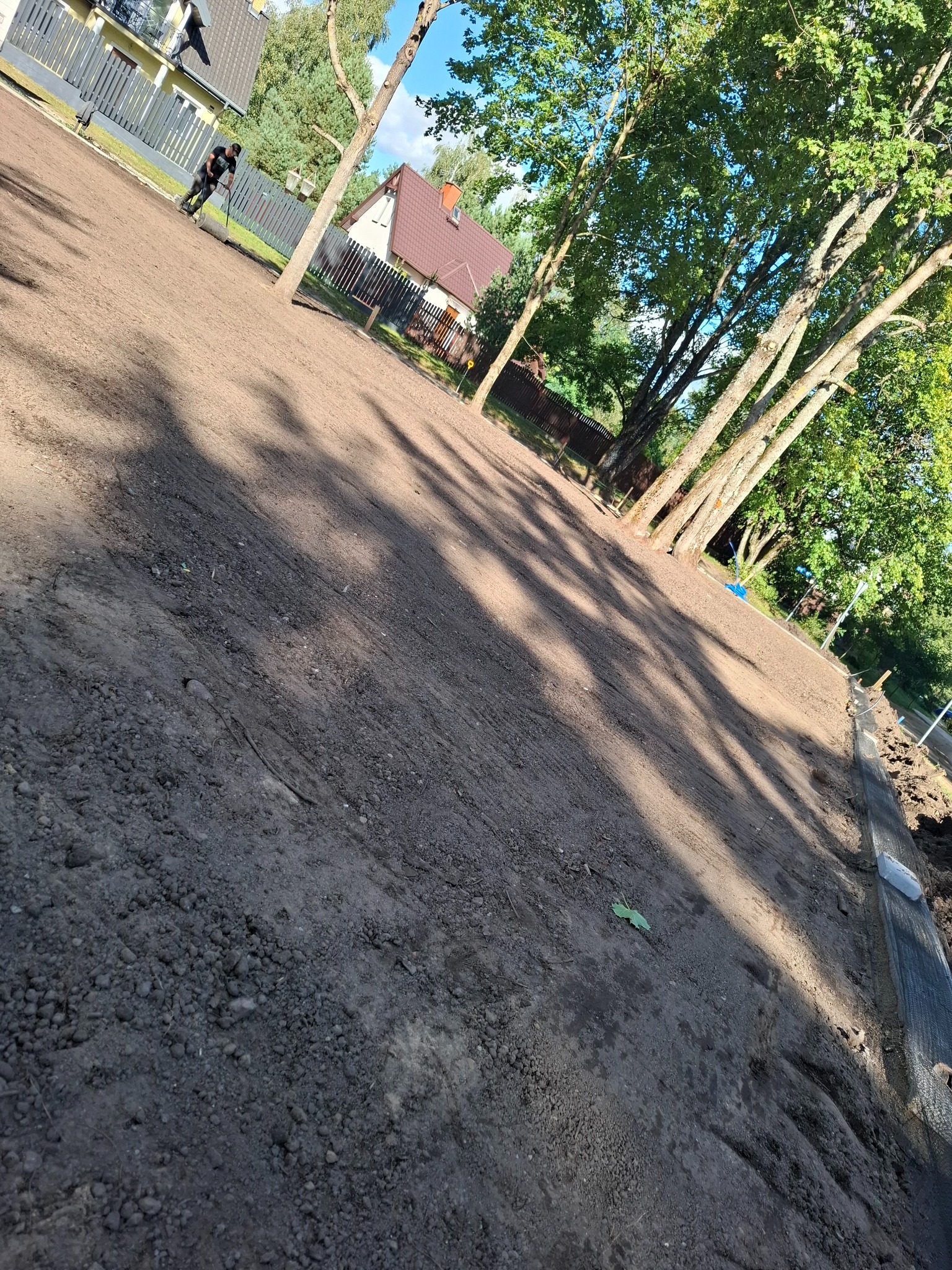 Przygotowany teren pod trawnik z rolki, wyrównana ziemia, widoczne drzewa i domy w tle, pracownik z wałem ogrodowym.