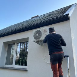 Marcin Frątczak - Montaż klimatyzatora na elewacji domu jednorodzinnego. Instalator na drabinie podłącza jednostkę zewnętrzną marki Konsi. Widok z dołu, błękitne niebo w tle.