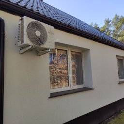 Marcin Frątczak - Boczny widok na zamontowaną jednostkę zewnętrzną klimatyzacji marki Konsi na elewacji domu z ciemnym dachem i rynnami. W oknach odbija się jesienny krajobraz.