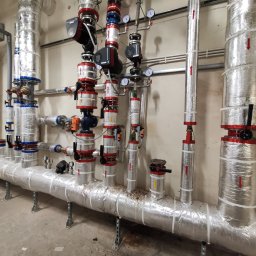 Marcin Frątczak - Izolowane rury instalacji grzewczej z zaworami i manometrami w pomieszczeniu technicznym. Rury owinięte folią aluminiową, z czerwonymi strzałkami wskazującymi kierunek przepływu.