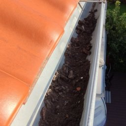 Piotrowski Mateusz - Zanieczyszczona rynna dachowa wypełniona ziemią, liśćmi i innymi zanieczyszczeniami, widok z góry, pomarańczowa dachówka w tle, fragment paneli słonecznych.