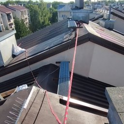 Piotrowski Mateusz - Montaż kolców przeciw ptakom na dachu z użyciem liny asekuracyjnej. Widok z góry na zabezpieczony fragment dachu i okoliczne budynki w słoneczny dzień.