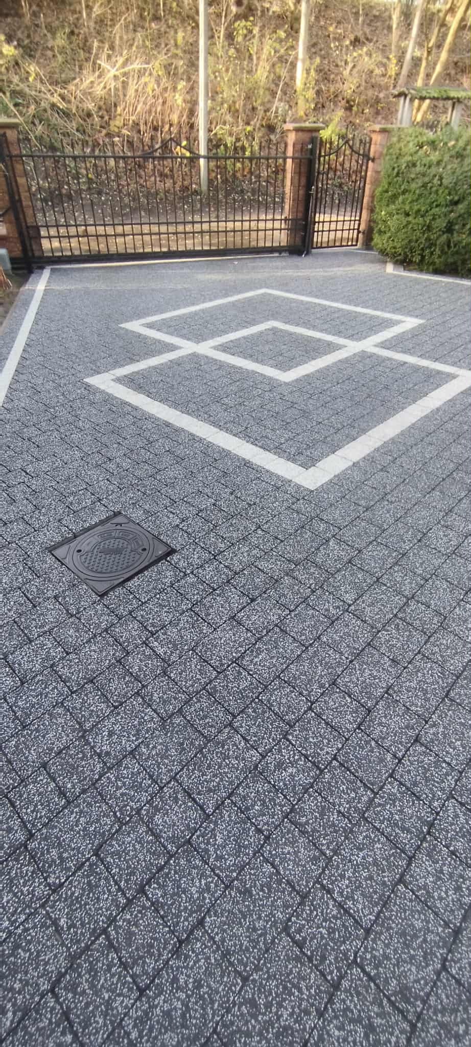 Szara kostka brukowa z białym wzorem i studzienką kanalizacyjną, brama w tle. Czysta nawierzchnia po umyciu, widok z góry.
