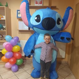 Two Friends Events - Chłopiec w okularach pozuje z animatorem przebranym za Stitcha, obok balony. Dekoracja sali z polską flagą w tle. Uśmiechnięty animator i dziecko.