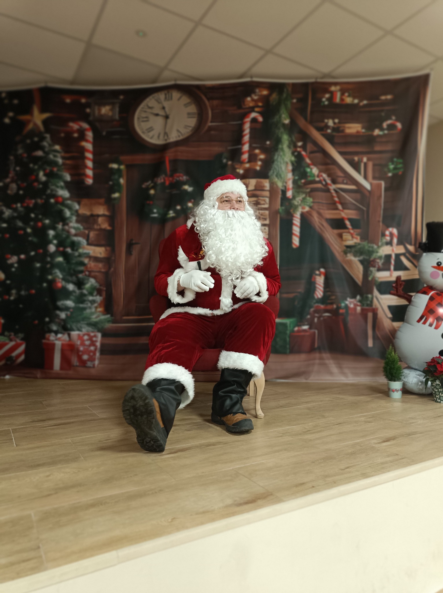 Uśmiechnięty Mikołaj w czerwonym stroju siedzi na krześle na tle świątecznej dekoracji w Poznaniu. Widoczny także dmuchany bałwan.