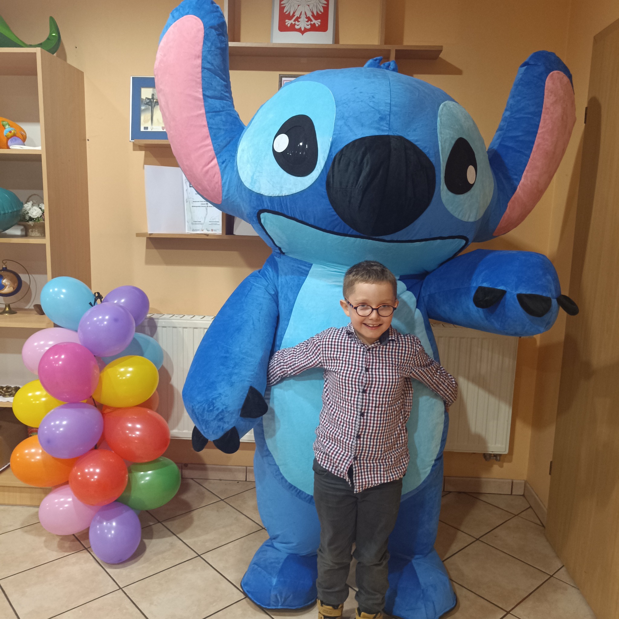 Chłopiec w okularach pozuje z animatorem przebranym za Stitcha, obok balony. Dekoracja sali z polską flagą w tle. Uśmiechnięty animator i dziecko.