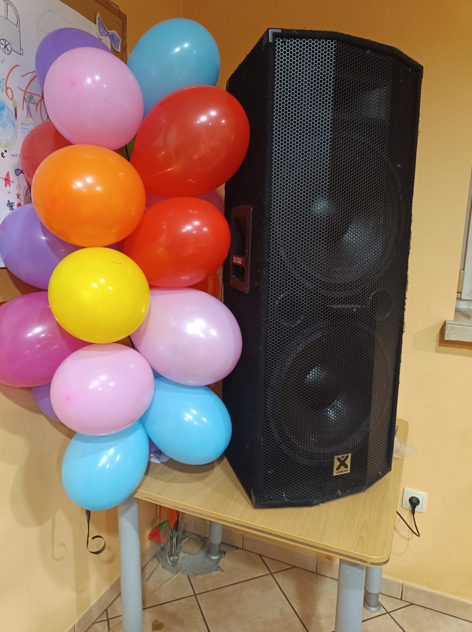 Kolorowe balony i czarny głośnik na stole, prawdopodobnie przygotowania do imprezy. Widoczne detale:  różne kolory balonów,  model głośnika.