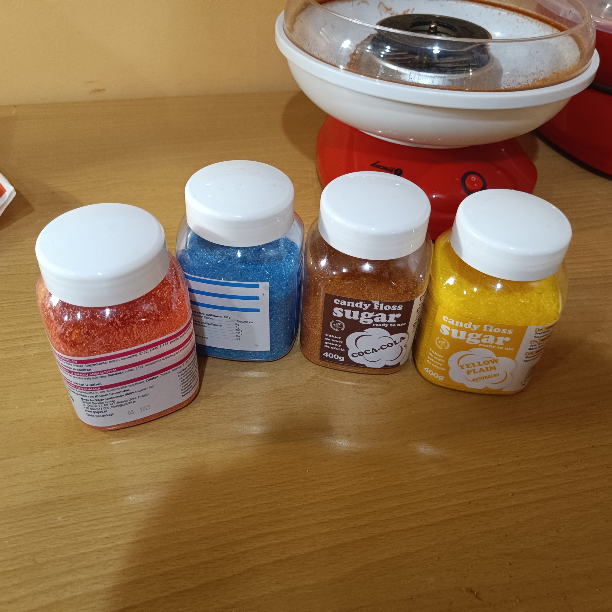 Kolorowe cukry do waty cukrowej (czerwony, niebieski, brązowy, żółty) w plastikowych pojemnikach, obok maszyna do waty cukrowej. Przygotowanie do imprezy.