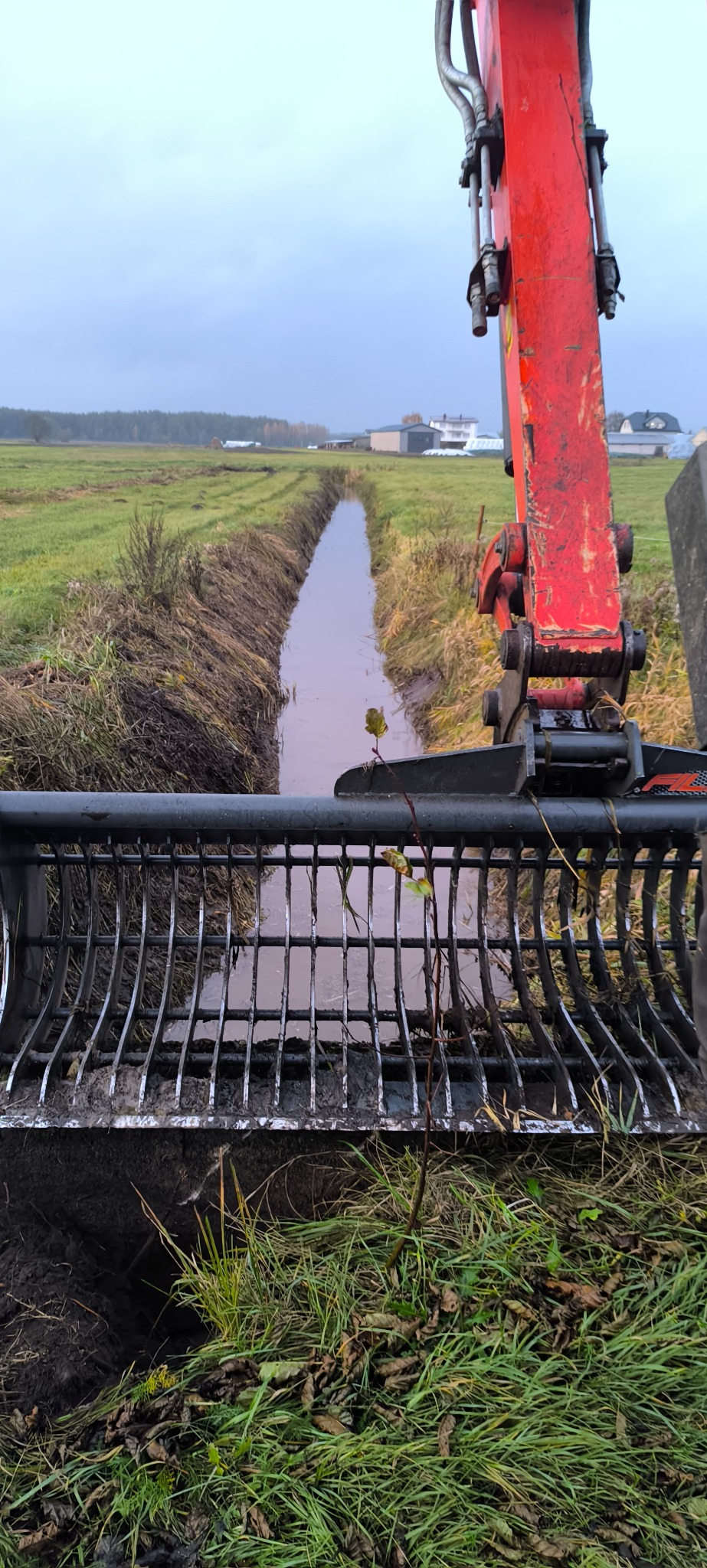 Wykopany rów melioracyjny widziany z perspektywy operatora koparki z łyżką ażurową. W tle łąka i zabudowania w pochmurny dzień.