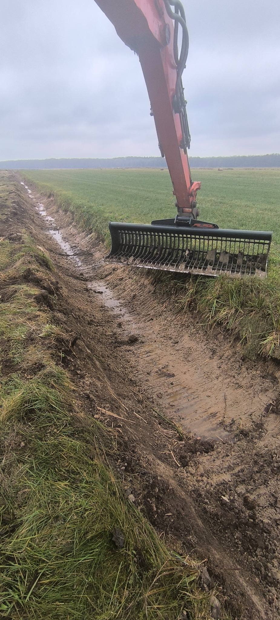 Czerwony traktor z czarną kratownicą kopie rów w ziemi na tle zielonego pola i pochmurnego nieba. Widok z bliska na wykopany rów wypełniony wodą.