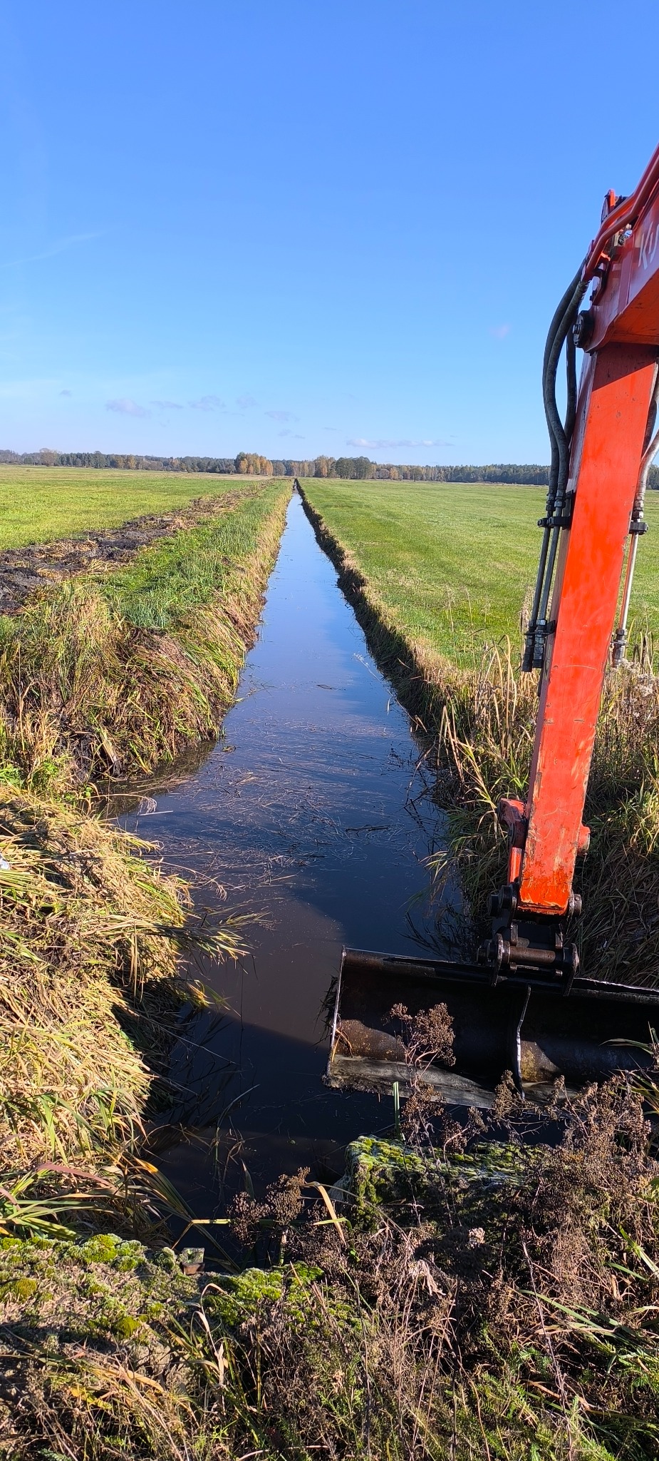 Pomarańczowa koparka przy rowie melioracyjnym na tle zielonego pola i błękitnego nieba. Rów wypełniony wodą, widoczne ślady świeżo wykonanych prac ziemnych.