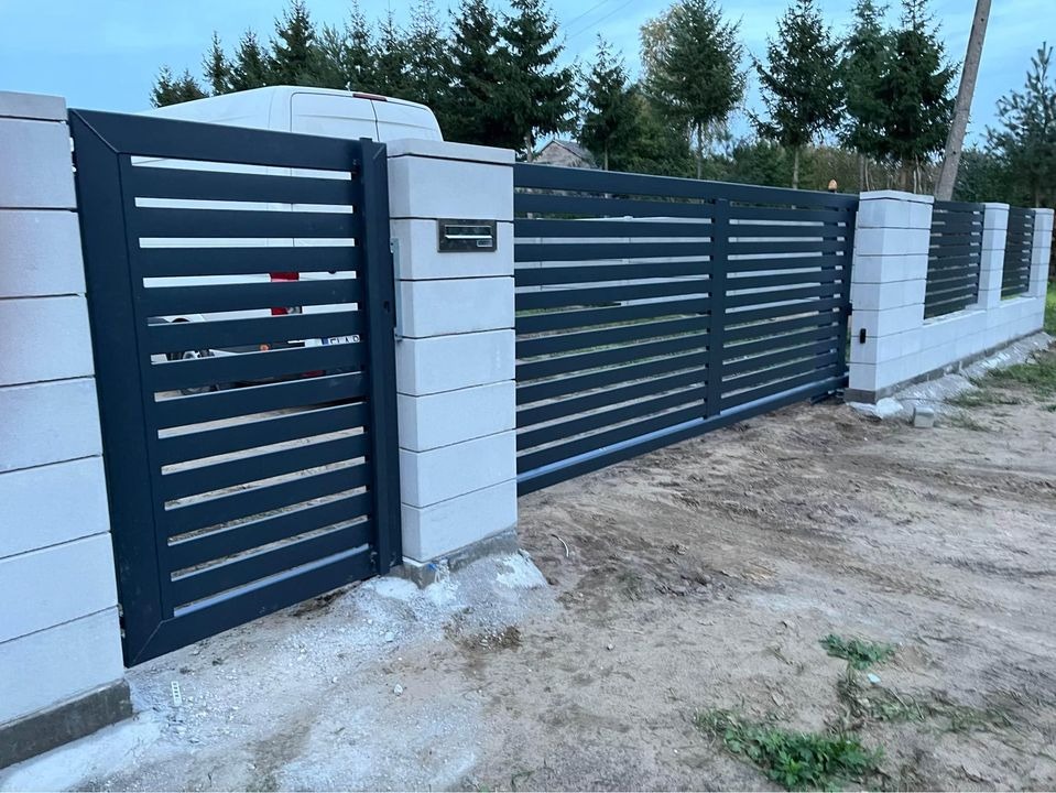 Nowoczesna, przesuwna brama metalowa z poziomymi szczeblami, osadzona na słupkach z szarego betonu. W tle widoczny biały bus i zielone drzewa.