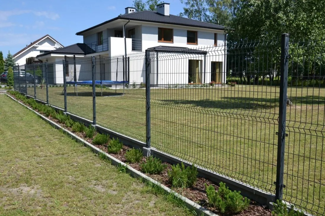 Nowoczesne ogrodzenie panelowe w kolorze grafitowym, oddzielające posesję z białym domem. Wzdłuż ogrodzenia rabata z roślinami i korą. Słoneczny dzień.