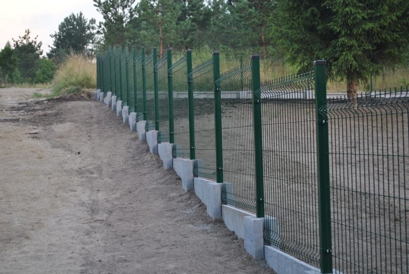 Zielone ogrodzenie panelowe z betonową podmurówką na nierównym terenie, widok z boku, w tle drzewa. Charakterystyczne mocowanie paneli do słupków.