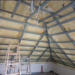 Usługi ogólnobudowlane Ad bud Adrian Kobędza - Skośny sufit poddasza z izolacją z pianki i metalową konstrukcją szkieletową, widoczne drabiny i tymczasowe oświetlenie. Etap prac wykończeniowych.