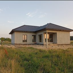 Usługi ogólnobudowlane Ad bud Adrian Kobędza - Surowy, jednokondygnacyjny dom z ciemną dachówką, w trakcie ocieplania, z widocznymi bloczkami fundamentowymi i elementami izolacji. W tle zieleń i błękitne niebo.