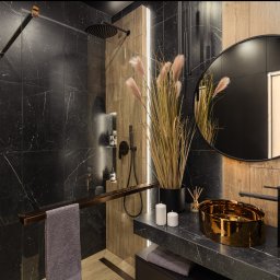 Usługi ogólnobudowlane Ad bud Adrian Kobędza - Nowoczesna łazienka z czarnymi płytkami imitującymi marmur, złotą umywalką i drewnianymi akcentami. Minimalistyczny design z okrągłym lustrem i dekoracyjnymi trawami.