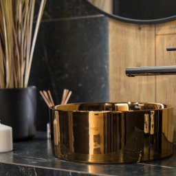 Usługi ogólnobudowlane Ad bud Adrian Kobędza - Złota umywalka nablatowa z czarną armaturą na blacie z ciemnego marmuru. W tle drewniana ściana i okrągłe lustro. Minimalistyczny wystrój łazienki.