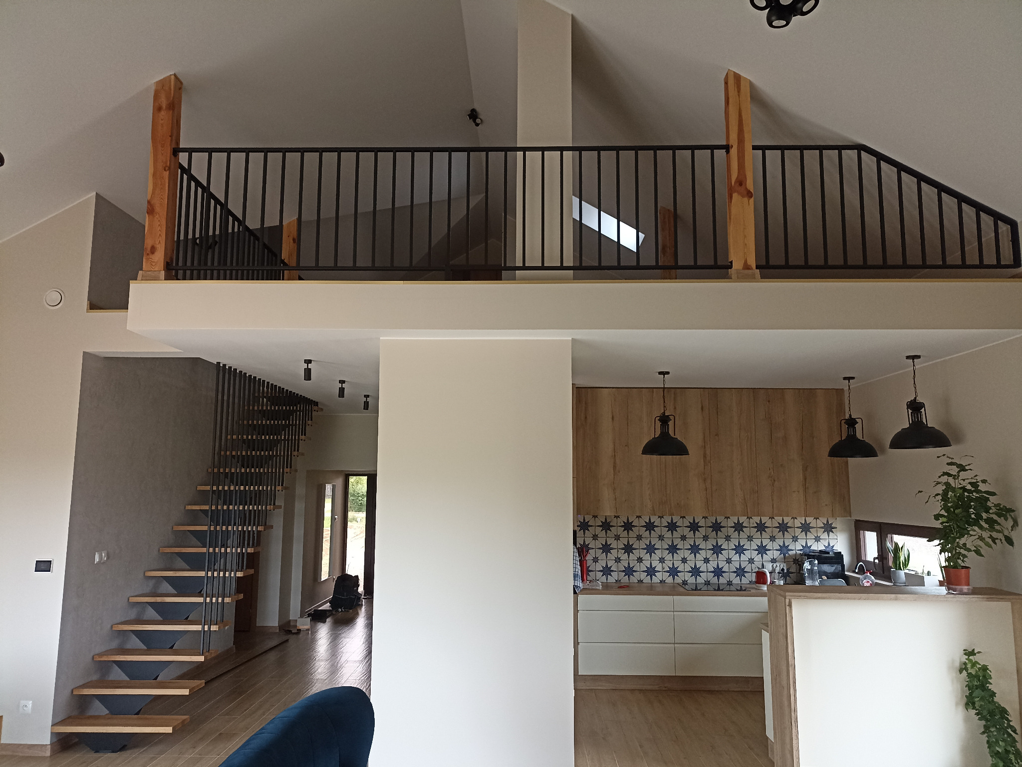Nowoczesne wnętrze domu z antresolą i schodami. Widoczne drewniane elementy, czarne balustrady i lampy. W tle kuchnia z niebieską mozaiką.