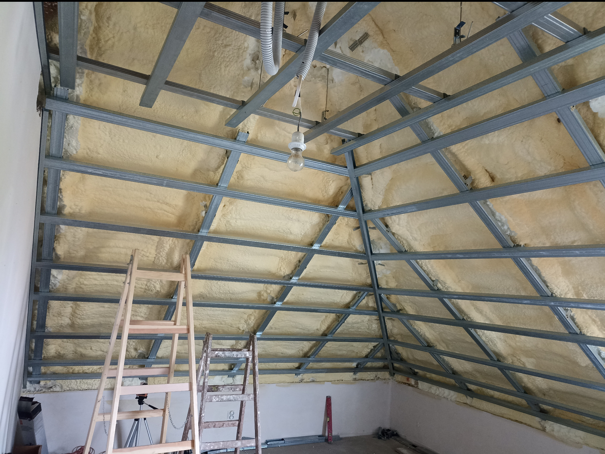 Skośny sufit poddasza z izolacją z pianki i metalową konstrukcją szkieletową, widoczne drabiny i tymczasowe oświetlenie. Etap prac wykończeniowych.