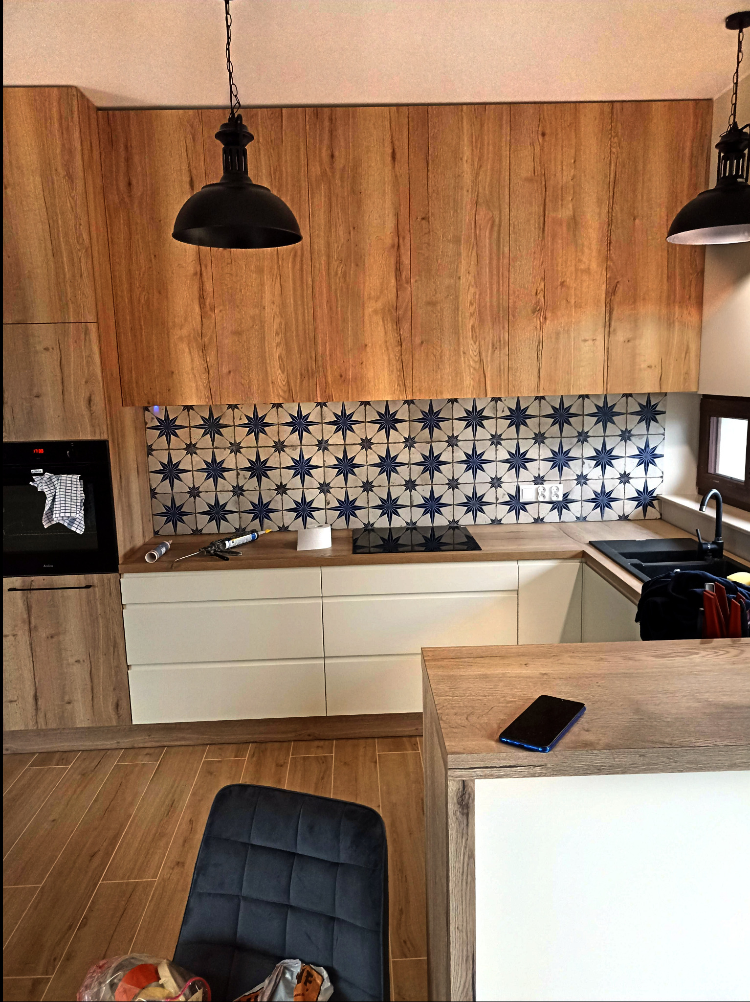 Nowoczesna kuchnia z drewnianymi frontami i płytkami w gwiazdy nad blatem. Widoczne narzędzia i telefon na blacie. Dwie czarne lampy wiszące nad wyspą.