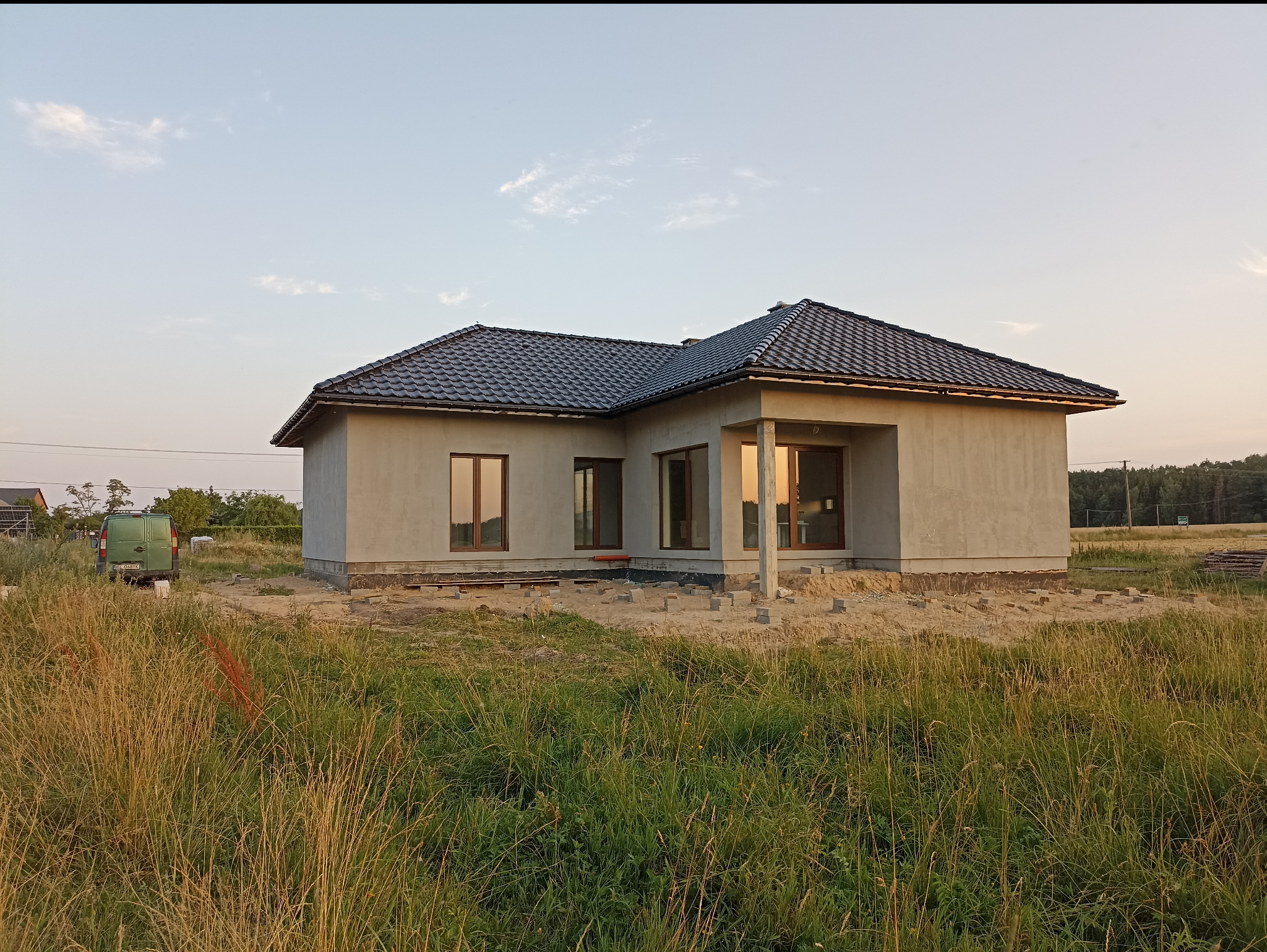 Surowy, jednokondygnacyjny dom z ciemną dachówką, w trakcie ocieplania, z widocznymi bloczkami fundamentowymi i elementami izolacji. W tle zieleń i błękitne niebo.
