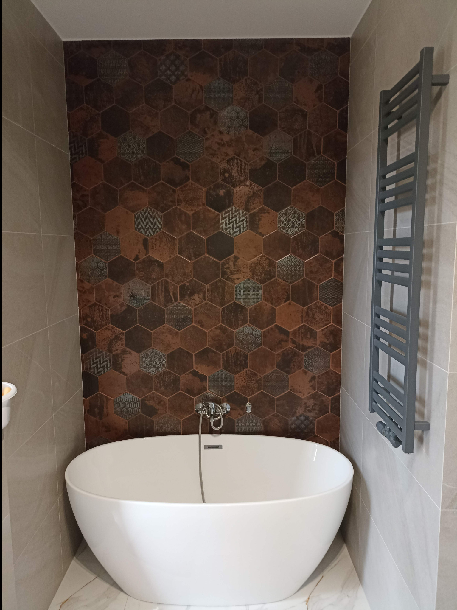 Nowoczesna łazienka z wolnostojącą wanną na marmurowej podłodze, ściana z heksagonalnych płytek w odcieniach brązu i szarości, grzejnik drabinkowy.