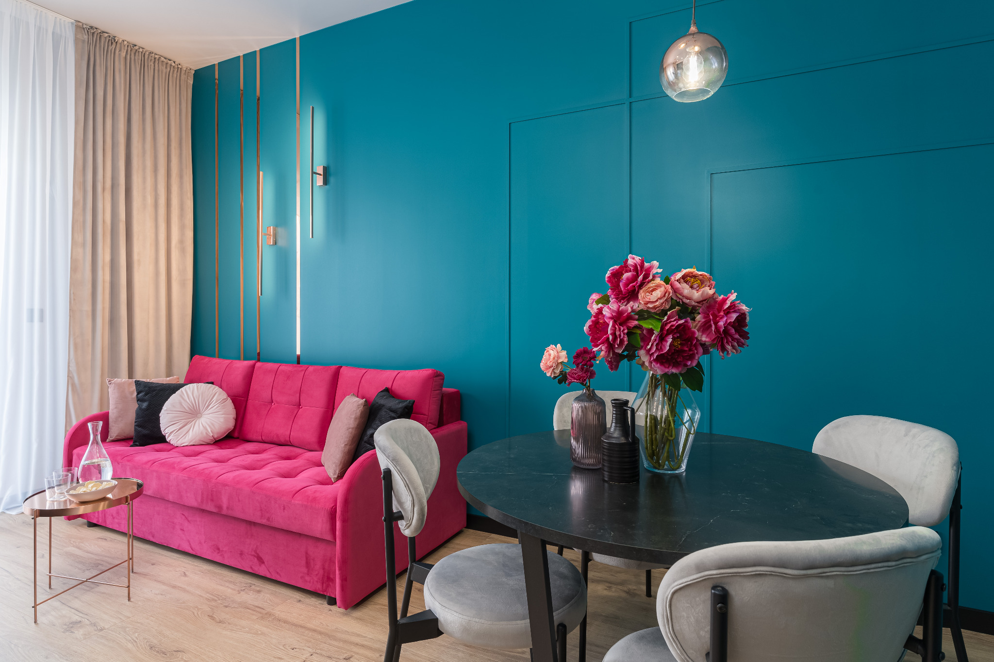 Turkusowa ściana z dekoracyjnymi listwami, różowa sofa i okrągły stół z kwiatami. Nowoczesne wnętrze z lampą wiszącą i drewnianą podłogą.