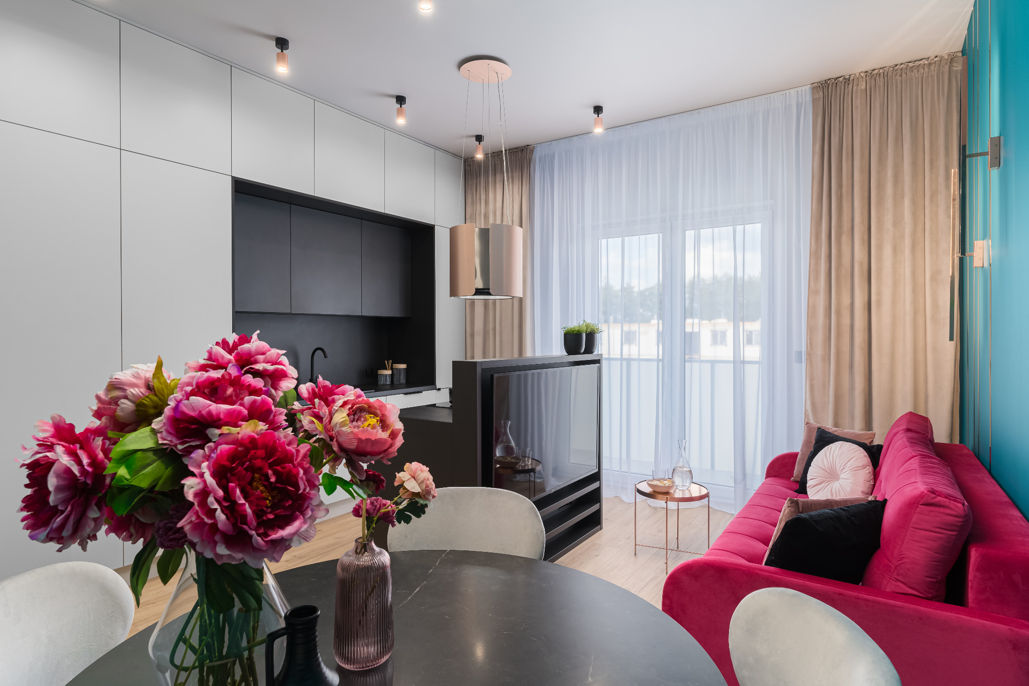 Nowoczesne wnętrze: czarno-biała kuchnia z wyspą, różowy aksamitny narożnik, okrągły stół z kwiatami. Minimalistyczny design z akcentami kolorystycznymi i naturalnym światłem.