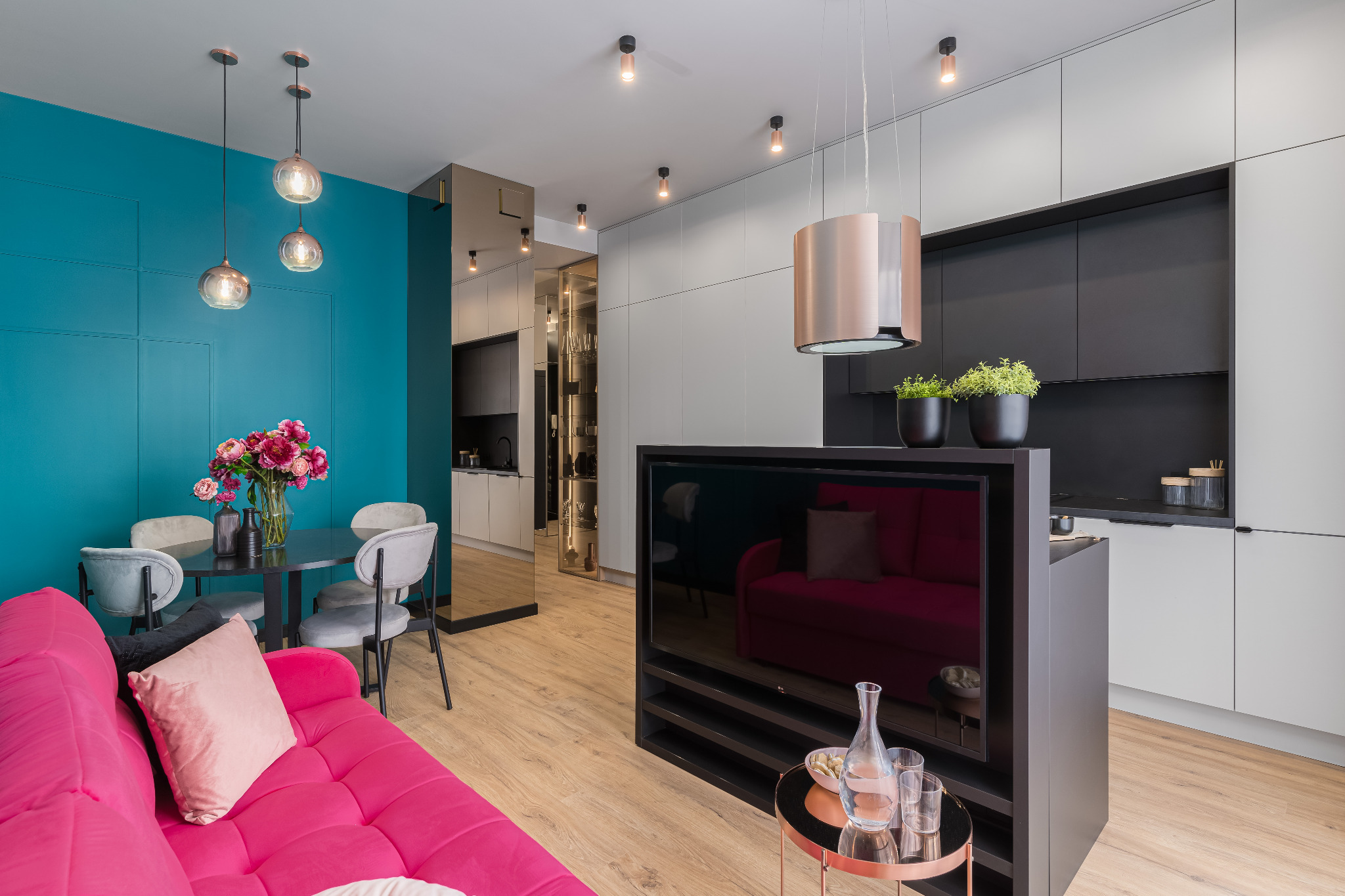 Nowoczesne wnętrze: turkusowa ściana z panelami, różowa sofa, stół z kwiatami i minimalistyczna kuchnia z czarnymi akcentami. Widok na salon z TV i dekoracjami.