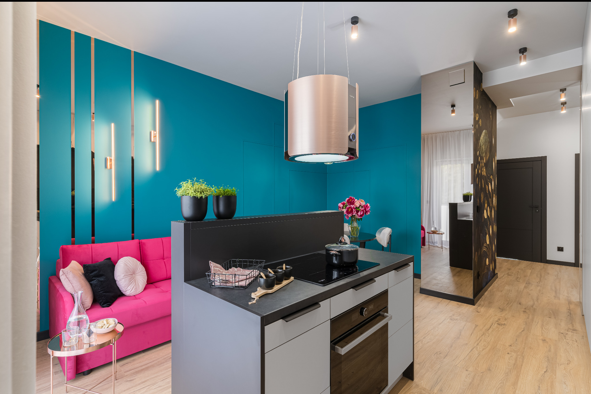 Nowoczesne wnętrze: turkusowa ściana z oświetleniem LED, różowa sofa, wyspa kuchenna z płytą i piekarnikiem, drewniana podłoga. Aranżacja z dbałością o detale.