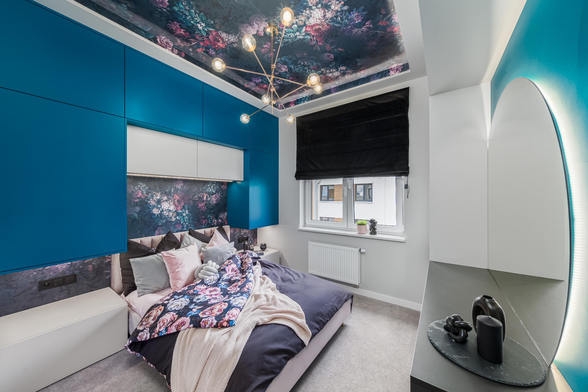 Nowoczesna sypialnia z niebieskimi szafkami, kwiatowym motywem na suficie i ścianie za łóżkiem. Centralnie umieszczone łóżko z dekoracyjną narzutą.