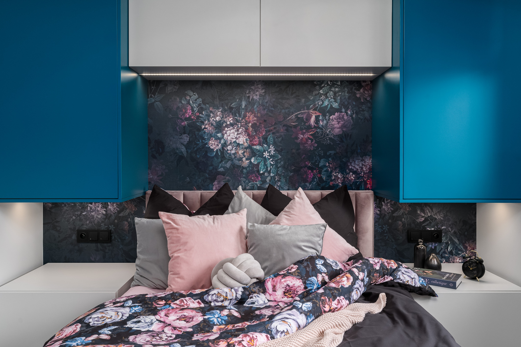 Nowoczesna sypialnia z tapetą w kwiaty, oświetleniem LED i niebieskimi szafkami. Widoczne dekoracyjne poduszki i narzuta w kwiatowy wzór. Minimalistyczny design.