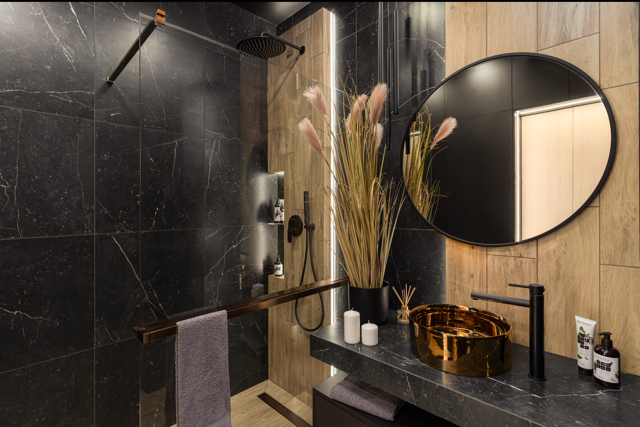 Nowoczesna łazienka z czarnymi płytkami imitującymi marmur, złotą umywalką i drewnianymi akcentami. Minimalistyczny design z okrągłym lustrem i dekoracyjnymi trawami.