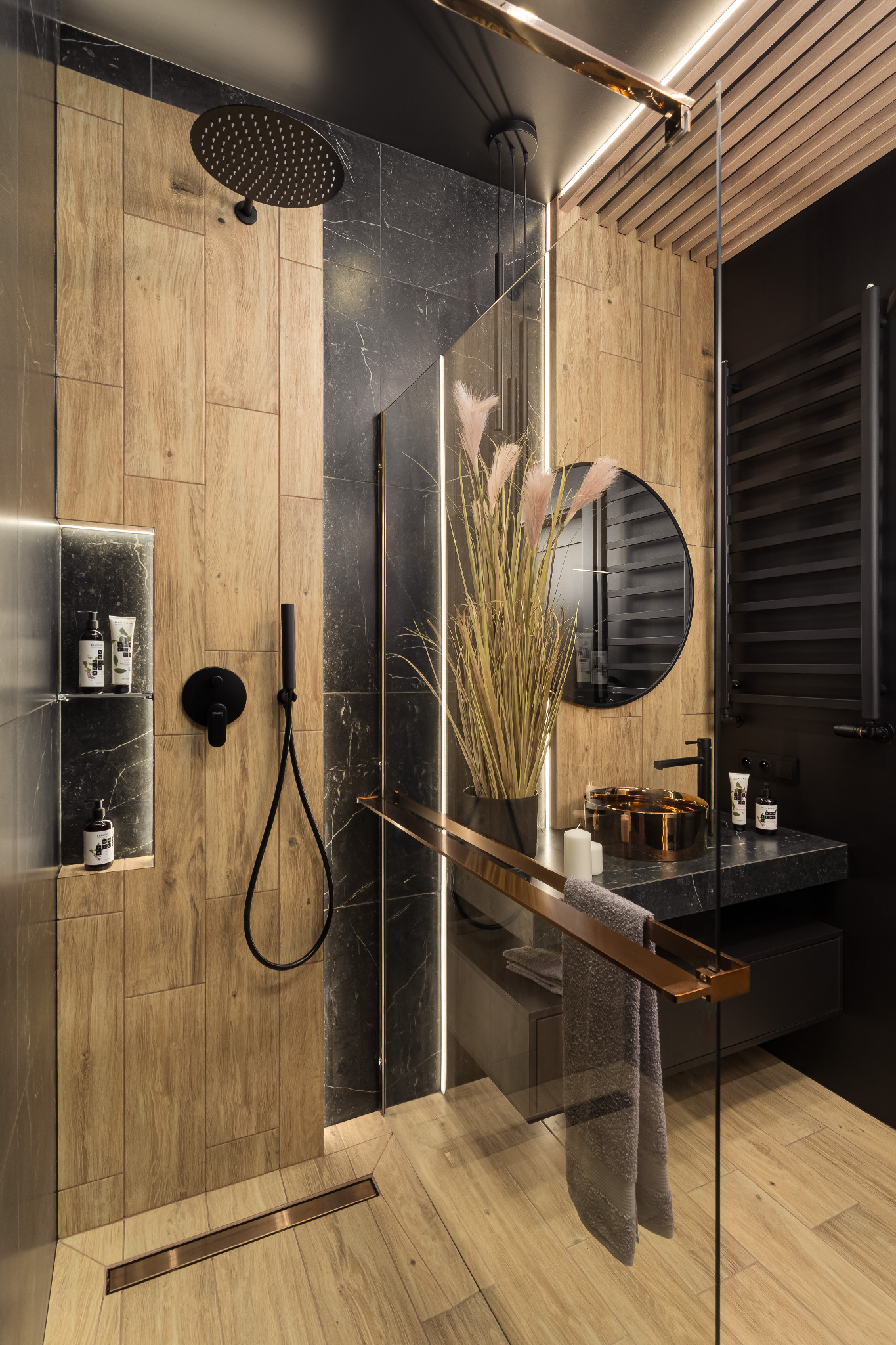 Nowoczesna łazienka z kabiną typu walk-in, drewnopodobnymi i marmurowymi płytkami, czarną armaturą i okrągłym lustrem. Minimalistyczny design z akcentami miedzi.