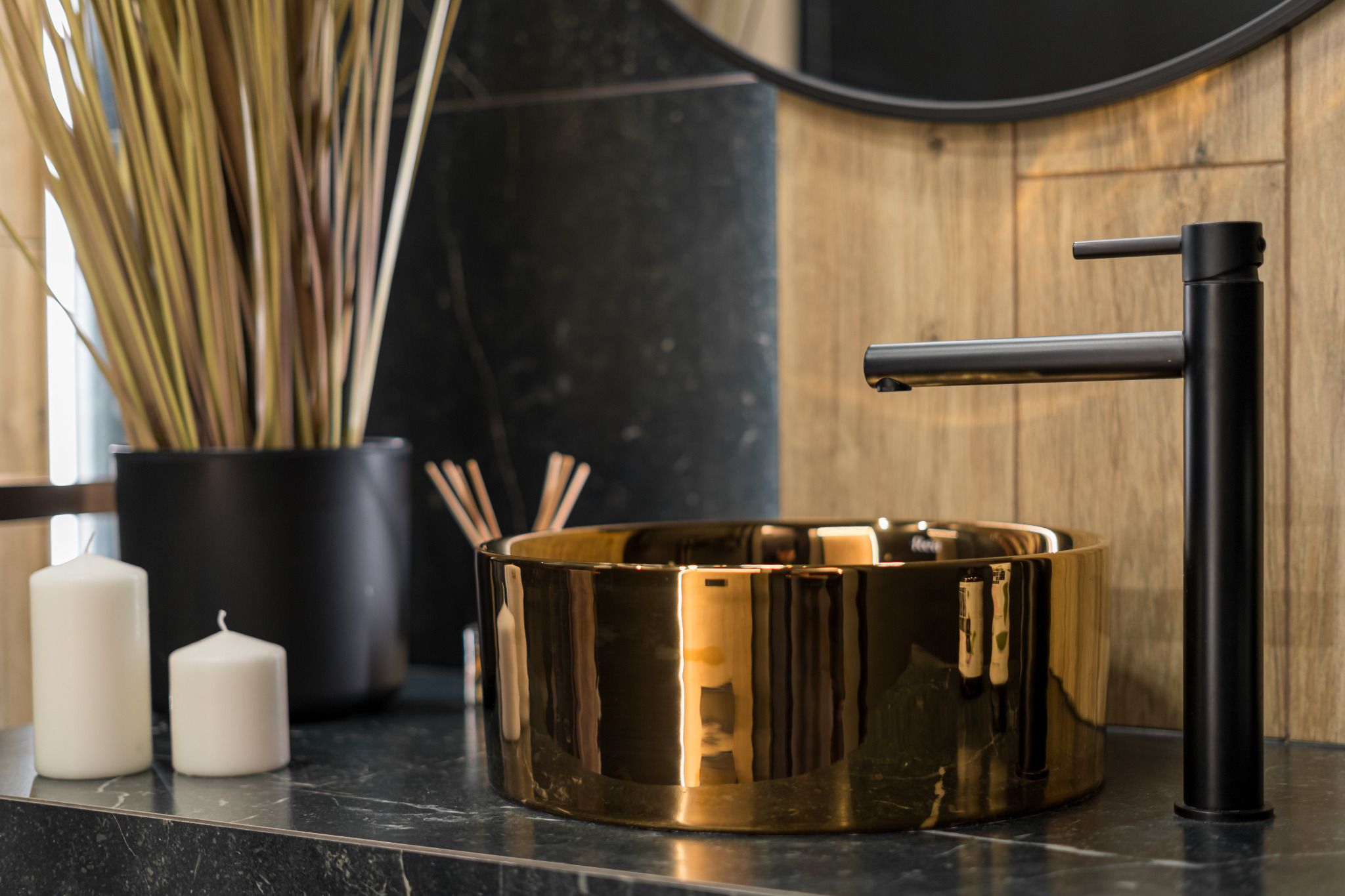 Złota umywalka nablatowa z czarną armaturą na blacie z ciemnego marmuru. W tle drewniana ściana i okrągłe lustro. Minimalistyczny wystrój łazienki.