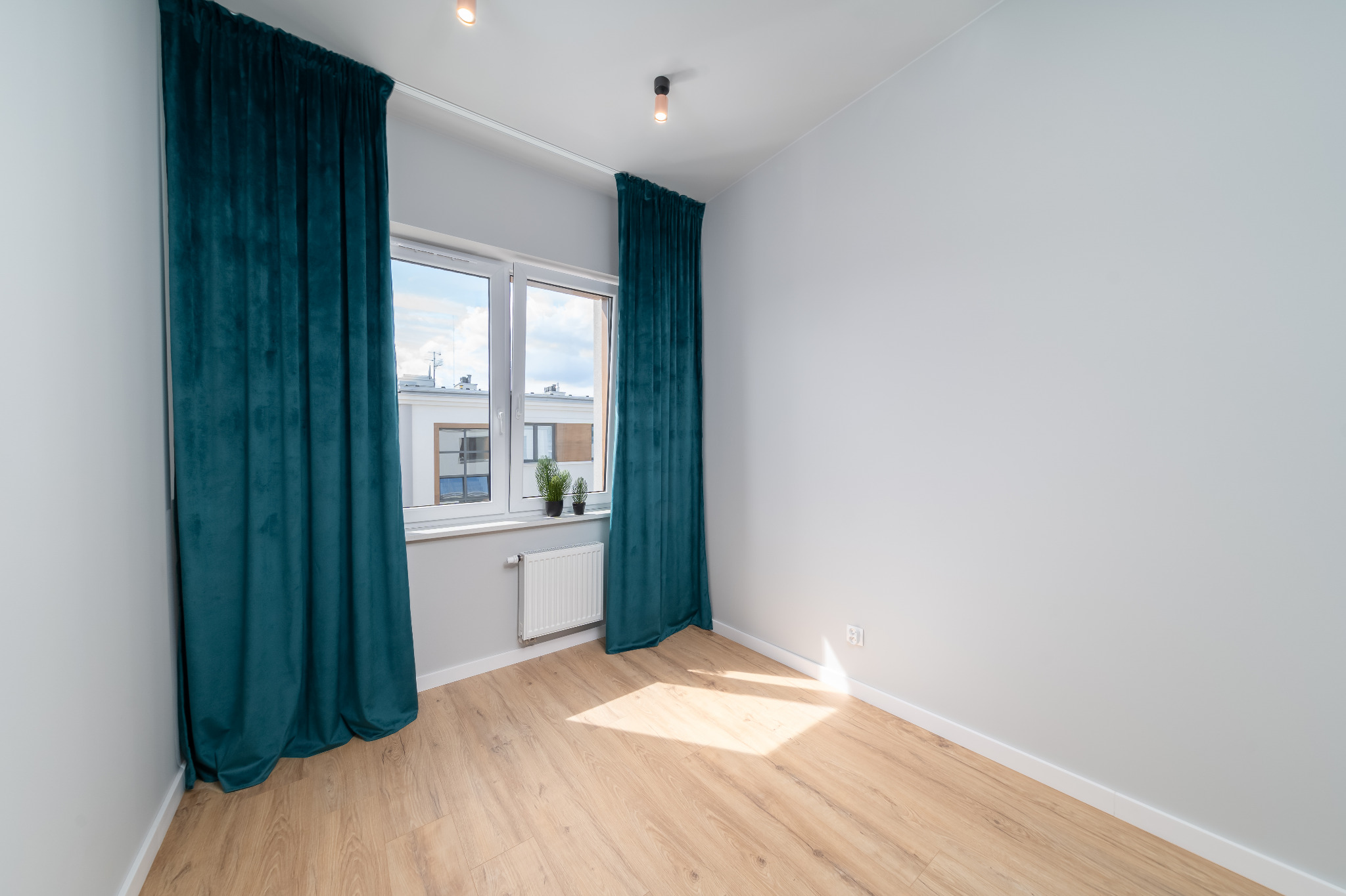 Jasne wnętrze pokoju z drewnianą podłogą, oknem z turkusowymi zasłonami i białymi ścianami. Minimalistyczny wystrój, widoczny grzejnik i kontakt.