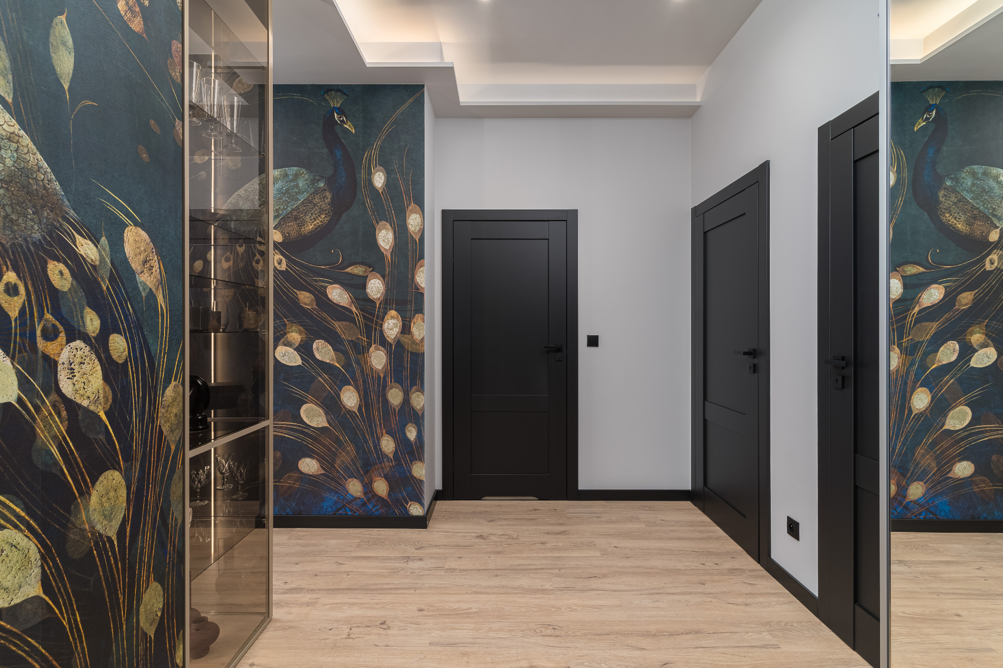Nowoczesny przedpokój z tapetą w pawie pióra, drewnianą podłogą i czarnymi drzwiami. Widoczna przeszklona szafa z oświetleniem.