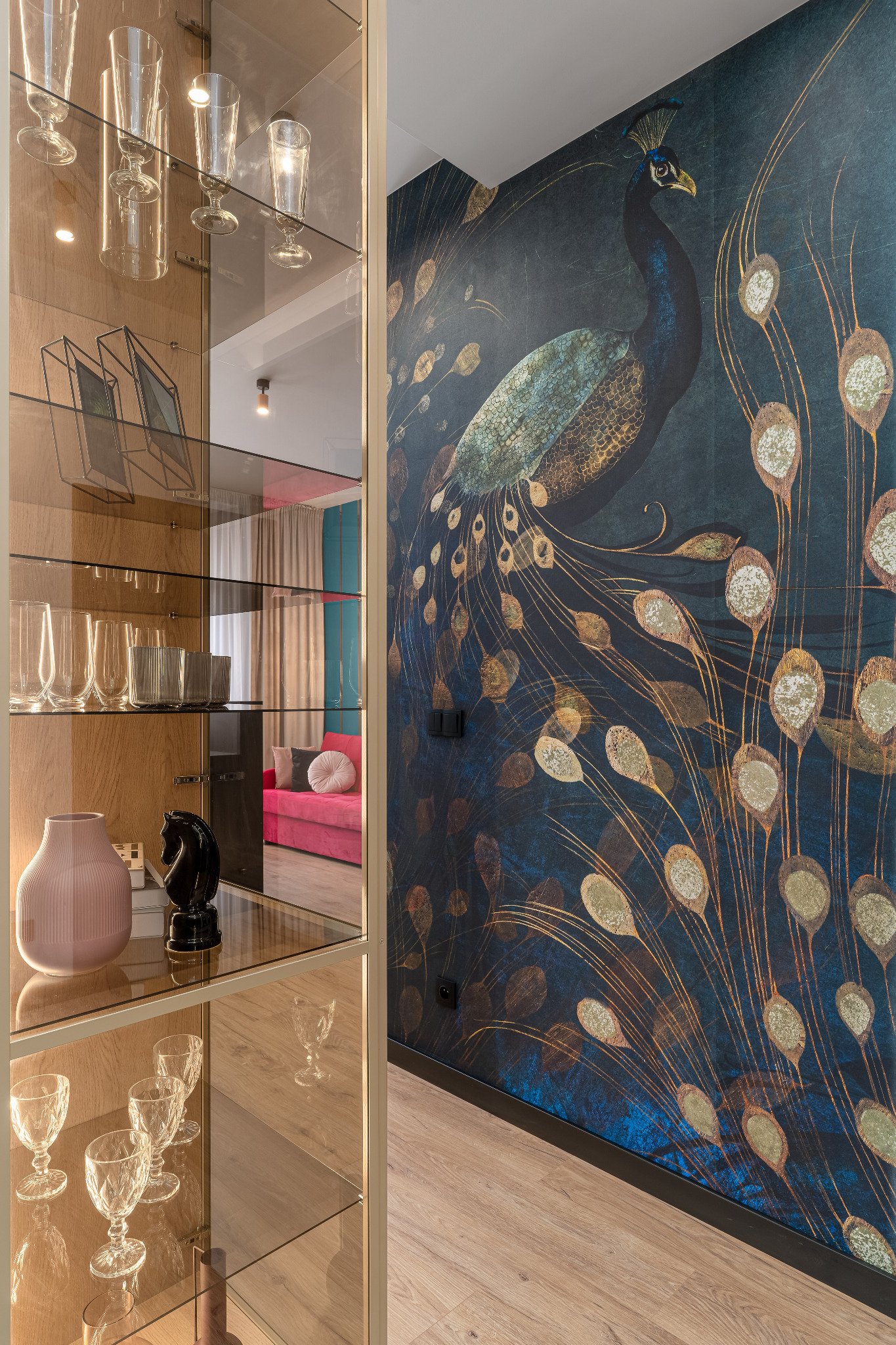 Stylowe wnętrze: szklana witryna z dekoracjami i tapeta z motywem pawia w odcieniach granatu i złota, widok na salon z różową sofą w tle.