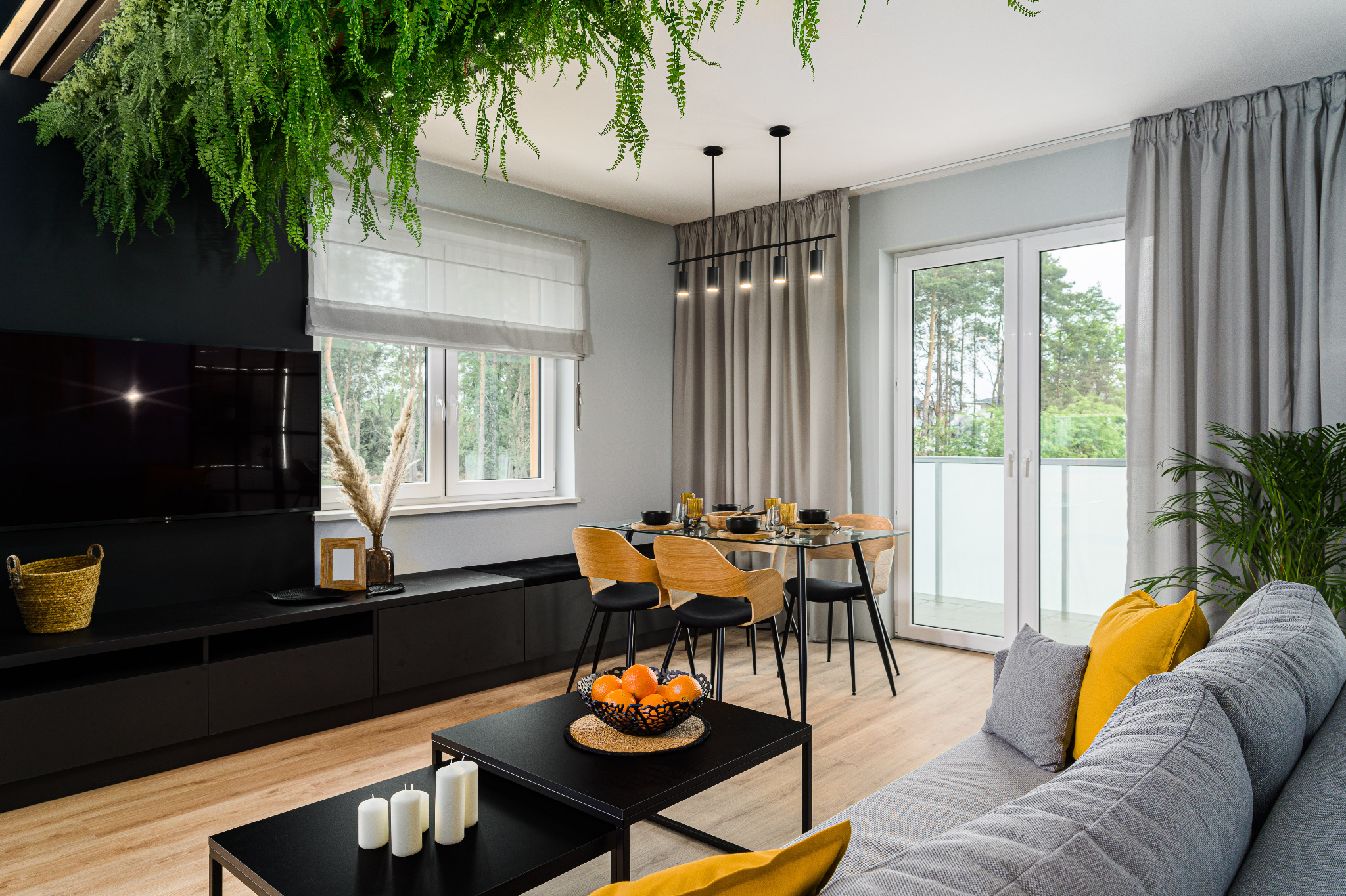 Nowoczesny salon z drewnianą podłogą, czarnymi meblami i szarą sofą z żółtymi poduszkami. Rośliny zwisają z sufitu, dodając naturalnego akcentu.